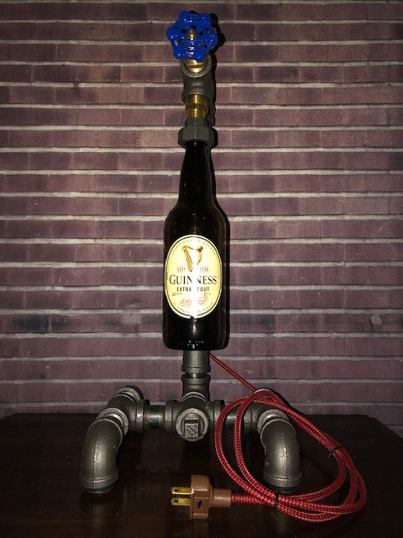 May include: A unique industrial-style lamp made from a Guinness beer bottle, metal pipes, and a blue valve. The lamp has a red power cord and a brown plug. The bottle features a gold and black label.