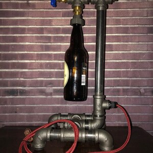 May include: A unique industrial-style lamp made from metal pipes, featuring a brown glass bottle as a central element. The lamp has a blue valve and a red electrical cord. The design is set against a brick wall background.