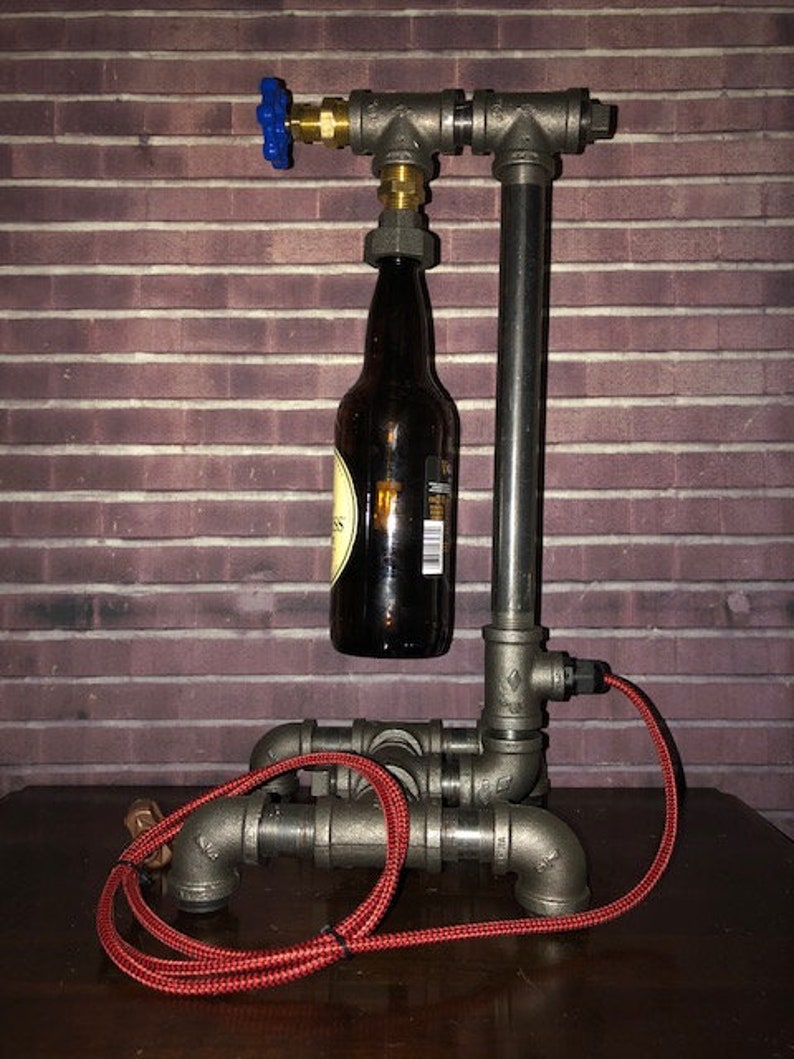 May include: A unique industrial-style lamp made from metal pipes, featuring a brown glass bottle as a central element. The lamp has a blue valve and a red electrical cord. The design is set against a brick wall background.