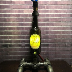 May include: A unique lamp made from a Guinness beer bottle, metal pipes, and a blue valve. The bottle is the central element, with the Guinness label visible. The lamp has a dark, industrial aesthetic, with a red wire.