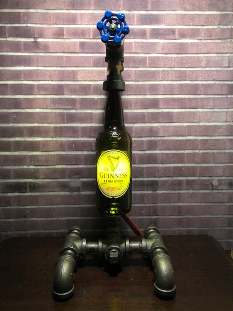 May include: A unique lamp made from a Guinness beer bottle, metal pipes, and a blue valve. The bottle is the central element, with the Guinness label visible. The lamp has a dark, industrial aesthetic, with a red wire.