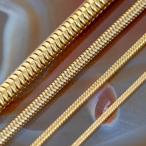 May include: Four gold-toned snake chains of varying thicknesses arranged diagonally. Each chain features a smooth, interlocking link design with a polished, reflective surface, showcasing a consistent metallic yellow hue across all four strands.