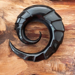 Könnte beinhalten: Ein schwarzer Ohr-Plug in Form eines spiralförmigen Horns mit segmentiertem, geschwungenem Design. Das Schmuckstück hat eine glatte, polierte Oberfläche, die sich zur Mitte der Spirale hin zu einer feinen Spitze verjüngt.