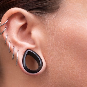Pode incluir: Close-up de uma orelha com múltiplos piercings. A orelha tem vários brincos de argola prateados e um grande plugue escuro em forma de gota. A pele é clara e texturizada.