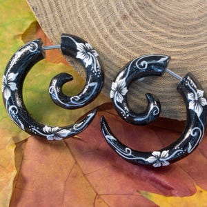 May include: A pair of black spiral-shaped earrings featuring a hand-painted white floral pattern with delicate swirling vine details. The earrings have a glossy finish and a thin metal post for insertion, creating a curved, tapered silhouette.