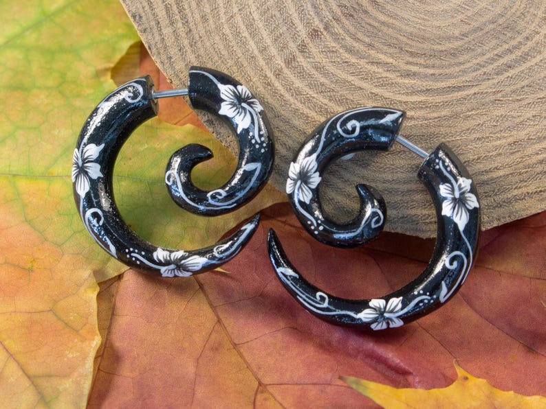 May include: A pair of black spiral-shaped earrings featuring a hand-painted white floral pattern with delicate swirling vine details. The earrings have a glossy finish and a thin metal post for insertion, creating a curved, tapered silhouette.