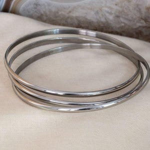 Pode incluir: Um conjunto de três pulseiras de metal prateado entrelaçadas com um acabamento liso e polido. As pulseiras circulares finas estão conectadas para formar uma unidade única, refletindo a luz nas suas superfícies arredondadas e simples.