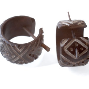 Puede incluir: Un par de pendientes de aro de madera marrón con un patrón geométrico blanco tallado en la superficie. Los pendientes tienen un simple poste de madera.