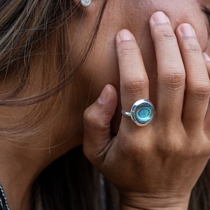 Puede incluir: Un anillo de plata con una incrustación de abulón azul y verde. El anillo está en el dedo de una persona.
