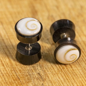 Può includere: Un paio di dilatatori per orecchie in legno nero e conchiglia bianca. I dilatatori hanno un design a spirale sul davanti.