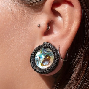 Può includere: Primo piano di un orecchio con un dilatatore per orecchie in abalone nero e iridescente a forma di serpente. Il dilatatore ha un anello d'argento nel lobo superiore.