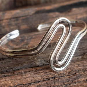 Può includere: Braccialetto d'argento con un design ondulato e astratto. Il braccialetto è realizzato in un unico pezzo di metallo che è piegato e attorcigliato in una forma unica.