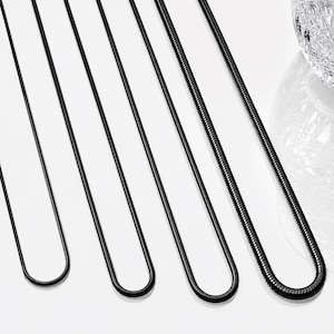 May include: A collection of black necklaces of varying lengths, displayed against a white background. The necklaces have a sleek, minimalist design, with a smooth, reflective surface. The chains are arranged in a row, showcasing their different sizes and styles.