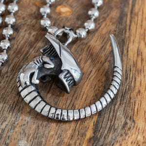 Könnte beinhalten: Ein silberfarbener Metallanhänger in Form einer Xenomorph-Kreatur mit länglichem Kopf und einem segmentierten Schwanz, der sich kreisförmig krümmt. Der Anhänger ist an einer silberfarbenen Kugelkette mit kleinen, runden, verbundenen Perlen befestigt.