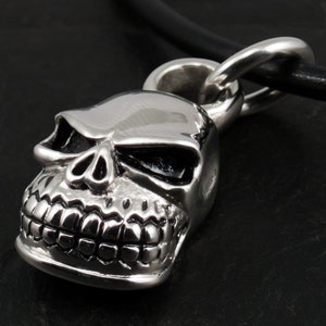 May include: A silver skull pendant with a detailed face and black eyes, hanging from a black leather cord.