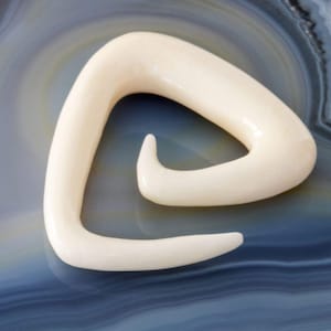 Op de afbeelding: Een witte spiraalvormige oorplug van bot, gevormd als een driehoek met een gebogen opening. De plug is op een witte en grijze achtergrond.