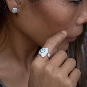 Puede incluir: Un anillo de plata con una incrustación de concha en espiral blanca y gris de forma triangular, junto con pendientes a juego. El conjunto de joyería presenta un acabado metálico pulido y un patrón de espiral natural en cada pieza.