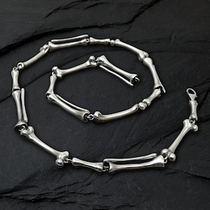 May include: A silver necklace with a bone design. The necklace is made up of multiple bone-shaped links joined together, creating a unique and edgy accessory. The clasp is visible at the end of the necklace.