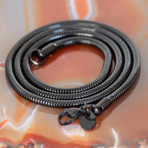 Op de afbeelding: Een opgerolde, zwarte ketting met een slangenkettingontwerp. De ketting heeft een karabijnsluiting en een klein, ovaal label. De sieraden worden gepresenteerd op een roodbruin oppervlak.