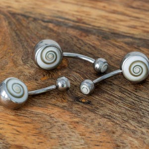 Può includere: Quattro gioielli per il corpo con un design a spirale. I gioielli hanno un metallo color argento e un motivo a spirale bianco e marrone. I pezzi sono disposti su una superficie di legno.