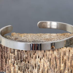 Pode incluir: Uma pulseira de metal cor de prata com um acabamento liso e polido. O interior da banda apresenta o texto gravado "flystyle" juntamente com um pequeno logótipo circular. A pulseira tem um design aberto, permitindo um ajuste fácil.