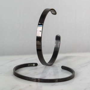 Op de afbeelding: Twee strakke, zwarte metalen armbanden met een gepolijste, reflecterende afwerking. De ene armband staat verticaal opgesteld, terwijl de andere horizontaal ligt. Beide hebben een eenvoudig, open ontwerp met een gladde, uniforme bandstructuur.