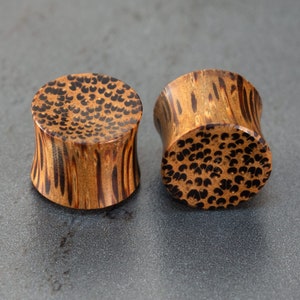 Puede incluir: Dos tapones para orejas de madera con un diseño de patrón marrón y negro. Los tapones son cilíndricos y tienen un acabado liso.