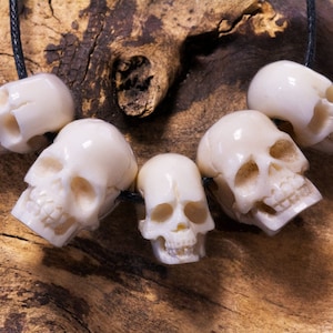 Könnte beinhalten: Eine Halskette mit sechs elfenbeinfarbenen Totenkopfperlen, die mit Gesichtsdetails geschnitzt sind und auf einer schwarzen Kordel aufgereiht sind. Die Halskette wird vor einem strukturierten Holzgrund präsentiert. Die Schädel sind etwa 2,5 cm hoch.