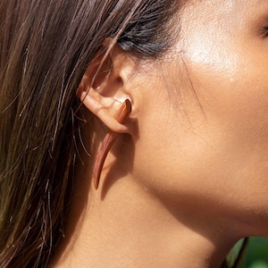 May include: A close-up of a person's ear with a long, wooden, curved earring. The earring is a natural wood colour and has a smooth finish.