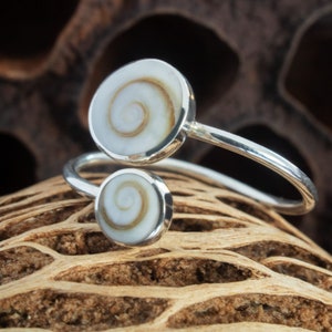 Puede incluir: Un anillo de plata con dos detalles de concha blanca y marrón. El anillo es abierto y tiene un diseño sencillo.