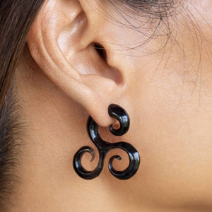 Puede incluir: Pendientes expansores de oreja negros en forma de espiral. Los expansores tienen un acabado brillante y un diseño de tres partes. La joya se lleva en la oreja y es de color oscuro y sólido. El diseño es un accesorio único y llamativo.