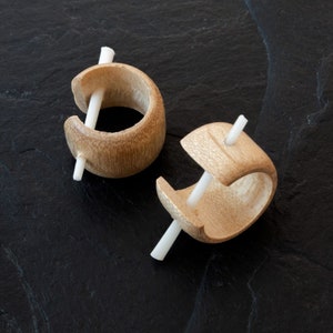 Pode incluir: Dois brincos de argola de madeira com uma haste de madeira branca no centro. Os brincos estão sobre uma superfície preta.