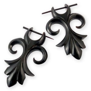 Könnte beinhalten: Ein Paar schwarze, geschnitzte Holzohrringe. Jeder Ohrring hat ein stilisiertes Blumendesign mit geschwungenen Linien und einer blattartigen Form am unteren Ende. Ein dunkler, gerader Stab ist durch die Oberseite jedes Ohrrings eingesetzt.