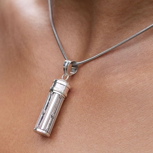 Könnte beinhalten: Eine silberne Halskette mit einem detaillierten, zylindrischen Anhänger, der sich öffnen lässt. Der Anhänger hat eine strukturierte Oberfläche und eine kleine Schlaufe oben.