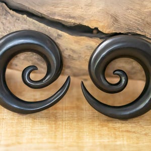 May include: A pair of black wood spiral ear gauges. The gauges are made from dark wood and have a smooth, polished finish.