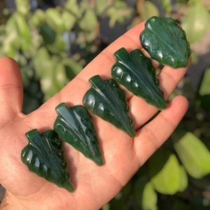May include: Five green jade leaf pendants with intricate carvings. The pendants are arranged in a row on a hand, with the largest pendant at the top and the smallest pendant at the bottom.