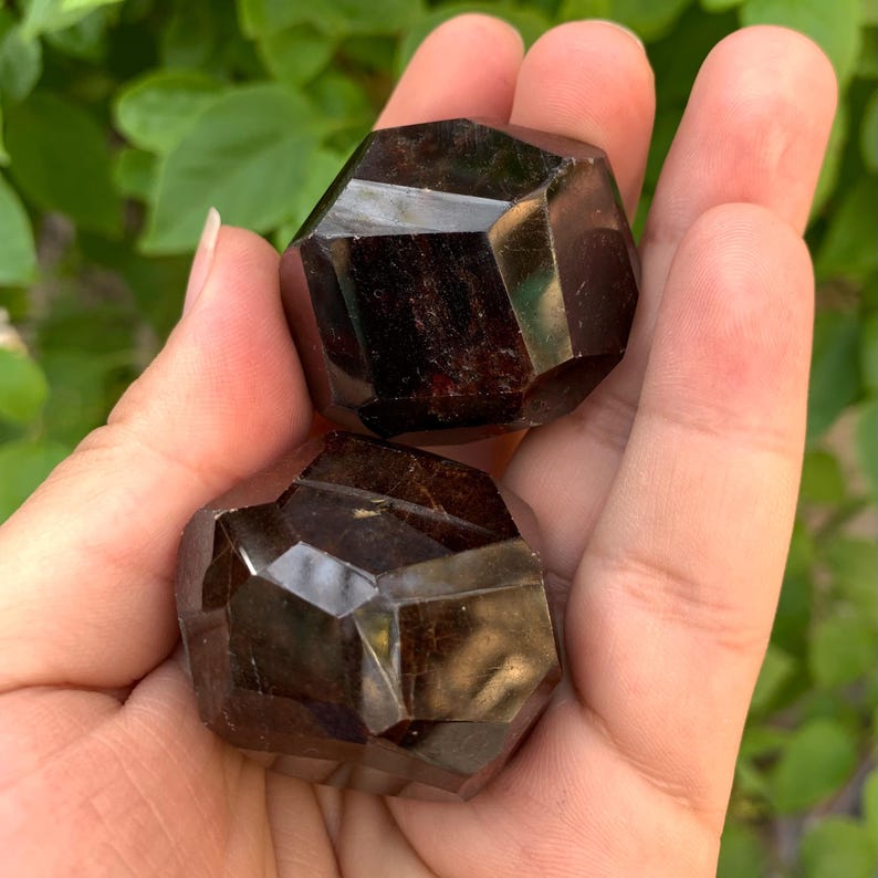 Puede incluir: Dos esferas de cristal de granate rojo oscuro, cada una con una forma geom&eacute;trica facetada. Las esferas se sostienen en una mano.