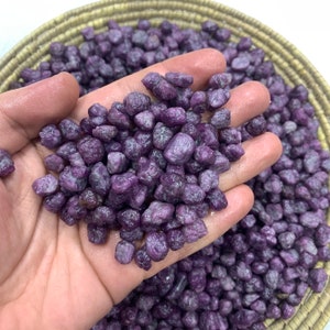 Puede incluir: Primer plano de una mano sosteniendo un montón de pequeñas piedras preciosas de color púrpura, talladas en bruto. Las piedras preciosas están en una cesta tejida.