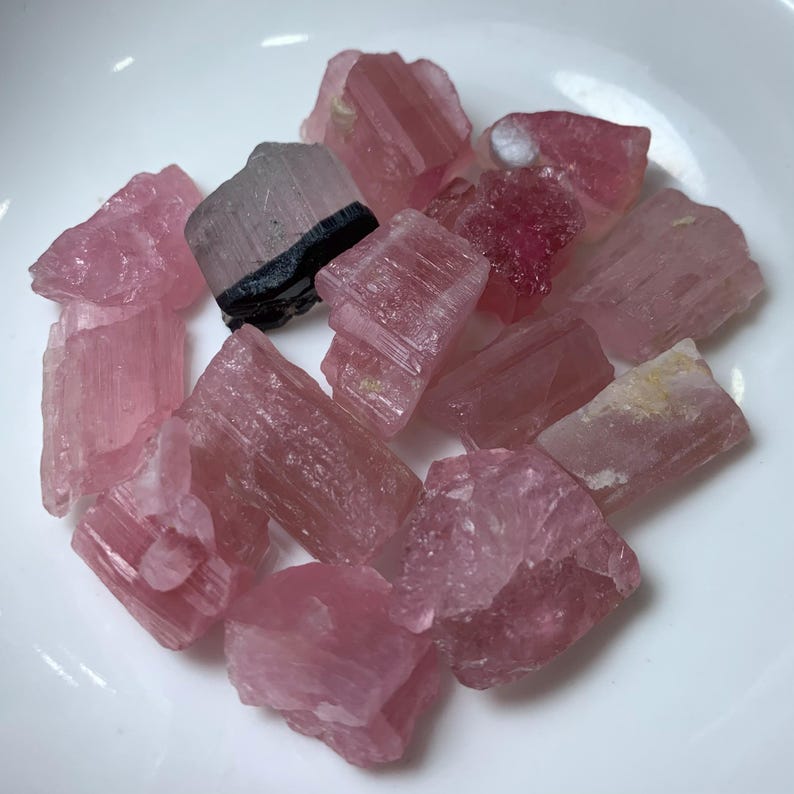 May include: A collection of pink and clear tourmaline crystals with black inclusions. The crystals are rough and have a natural, unpolished look.