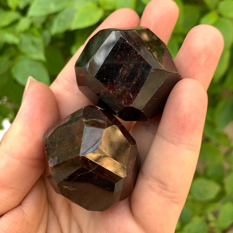 Puede incluir: Dos cristales de granate rojo oscuro, facetados, en forma de cubo. Los cristales se sostienen en una mano.