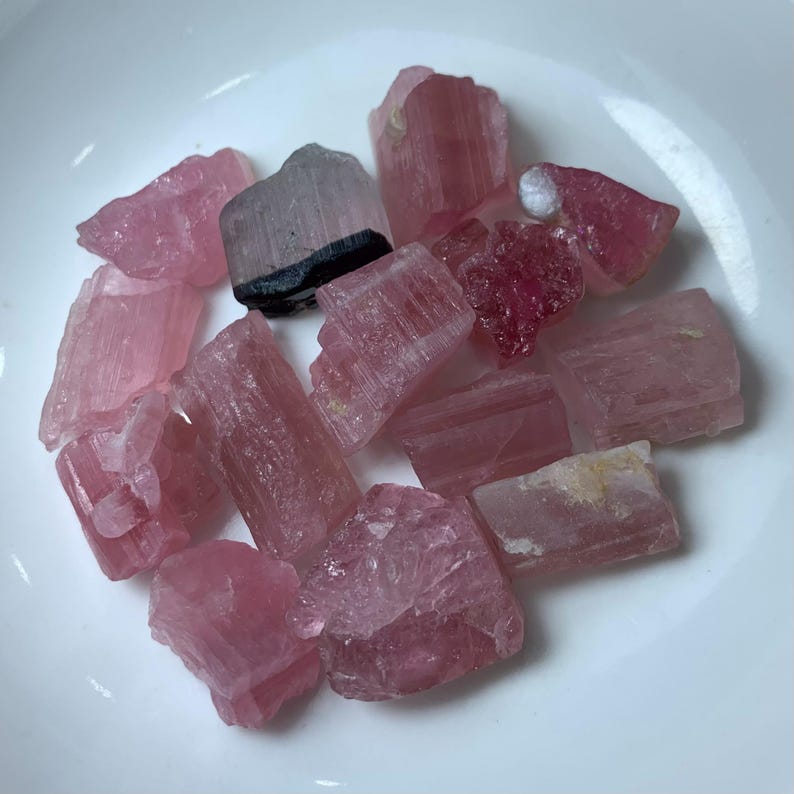 May include: A collection of pink and black tourmaline crystals. The crystals are raw and have a natural, rough texture. They are a variety of pink, from light to dark, and some have black streaks.