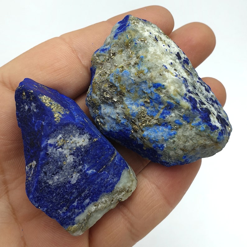 May include: Two rough, natural, blue lapis lazuli stones with gold pyrite inclusions. The stones are in a hand, showing their size and texture.