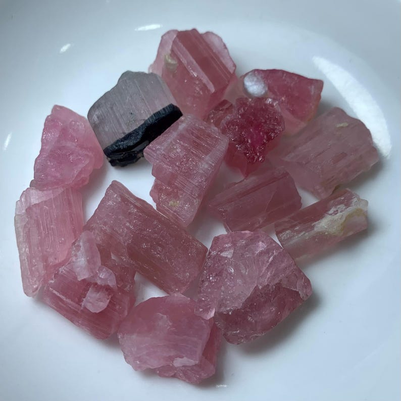 May include: A collection of pink and clear tourmaline crystals in various shapes and sizes. The crystals are arranged on a white surface.