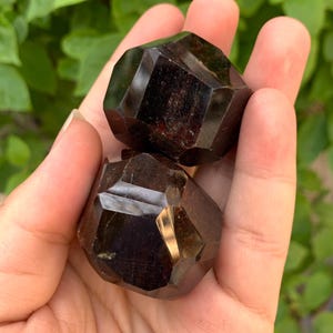 Puede incluir: Dos cristales de granate rojo oscuro, cada uno con una forma geom&eacute;trica facetada. Los cristales se sostienen en una mano, mostrando su tama&ntilde;o y detalles intrincados.