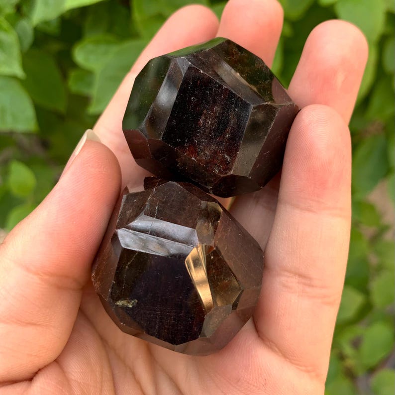 Puede incluir: Dos cristales de granate rojo oscuro, cada uno con una forma geom&eacute;trica facetada. Los cristales se sostienen en una mano, mostrando su tama&ntilde;o y detalles intrincados.