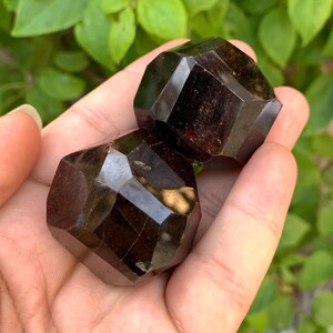 Puede incluir: Dos esferas de piedra preciosa facetadas de color rojo oscuro. Las esferas son lisas y pulidas, con un ligero brillo. Las esferas se sostienen en una mano, con el fondo desenfocado.