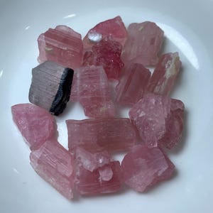 May include: A collection of pink and black tourmaline crystals. The crystals are rough and have a natural, unpolished look. They are a variety of pink and have a striated texture.