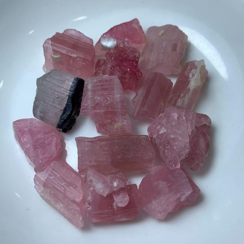 May include: A collection of pink and black tourmaline crystals. The crystals are rough and have a natural, unpolished look. They are a variety of pink and have a striated texture.