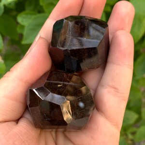 Puede incluir: Dos cristales de piedra preciosa de color marr&oacute;n oscuro facetados. Los cristales tienen la forma de un cubo con bordes redondeados. Los cristales se sostienen en una mano.
