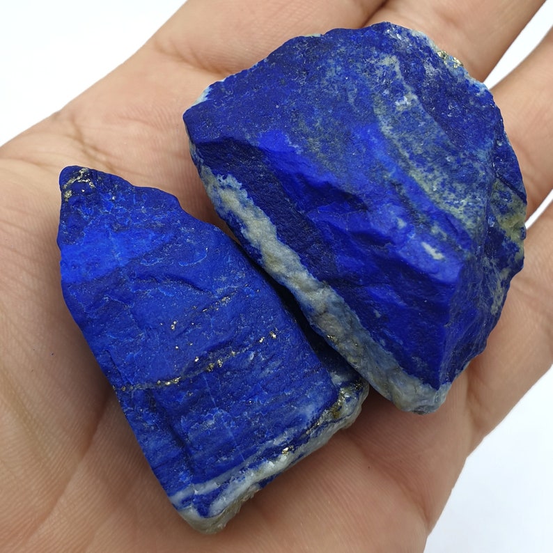 May include: Two rough, natural pieces of lapis lazuli, a deep blue gemstone with white and gold veins. The stones are irregular in shape and have a rough, unpolished texture.